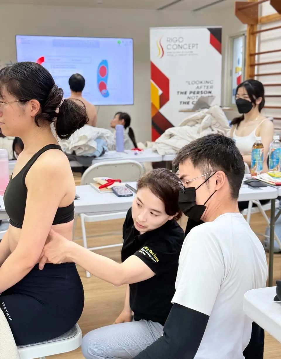 課堂實作：講師以手觸診坐姿示範學員的腰骨盆區，一旁學員近距離觀察手法，背景為投影講義與 Rigo Concept 展架